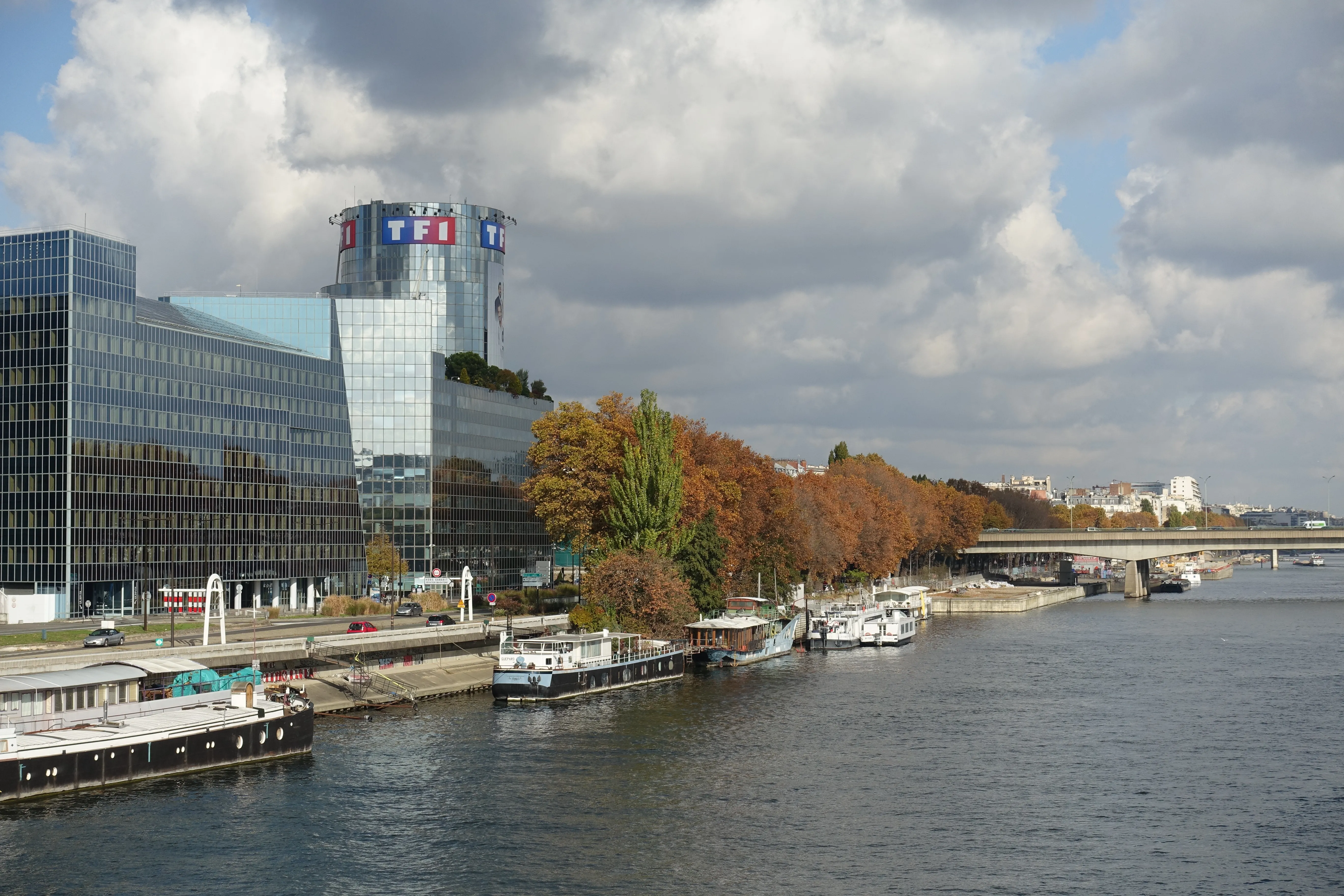 Tour TF1 vitrée au logo rouge à Boulogne-Billancourt, vue depuis le pont d'Issy-les-Moulineaux, avec péniches alignées le long du quai et arbres aux couleurs d'automne au premier plan, sous un ciel nuageux.