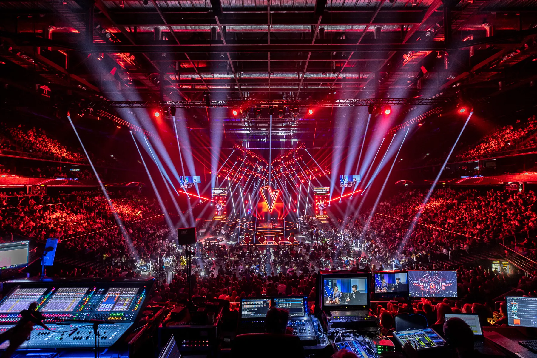 Vue depuis la régie technique d'une arène en finale du télé-crochet The Voice : public lointain, scène centrale éclairée en rouge, logo en V lumineux.